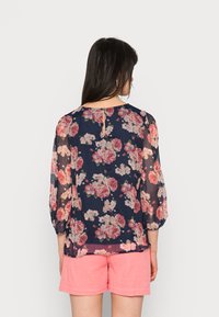 Blusa fluida azul marinho com padrão floral rosa, tecido transparente, mangas elásticas e fecho em gota nas costas. Combinada com calções rosa claro.