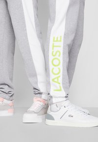 Grijze joggers met witte zijpanelen en groene "LACOSTE" tekst, gecombineerd met wit en grijze sneakers met roze accenten en branding.