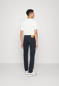 Vaqueros azul oscuro con un parche de cuero y una etiqueta roja en la parte trasera, combinados con una camiseta blanca básica y zapatillas atléticas grises.