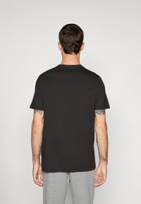 Mand med kort mørkt hår iført en ensfarvet sort T-shirt og grå bukser, stående med ryggen til foran en hvid baggrund.