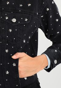 Černá džínová bunda s ozdobami ve tvaru stříbrných hvězd a hroty. Zapínání na knoflíky, viditelné kapsy a detaily na manžetách. Texturovaná látka.