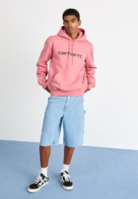 Pink hooded sweatshirt with a front pocket and black logo, paired with light blue denim shorts and black sneakers with white accents.
