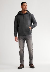 Sudadera gris con cremallera y capucha ajustable, dos bolsillos frontales, combinada con jeans gris claro y botas marrón oscuro, diseño minimalista.