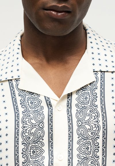 Κοντινό πλάνο ενός ατόμου που φοράει μια λευκή μπλούζα με μαύρα περίπλοκα μοτίβα πεϊσλί και γεωμετρικά σχέδια και ανοιχτό γιακά.