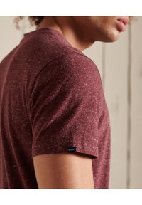 Burgunderfarbiges, strukturiertes T-Shirt mit kurzen Ärmeln, das ein kleines blaues Etikett trägt und auf der Schulter eines Modells vor einem hellen Hintergrund präsentiert wird.