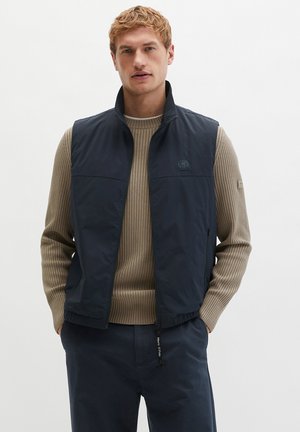 Mann trägt einen beige gerippten Pullover und eine aufgezippte blaue Weste, mit den Händen in den Taschen der blauen Hose vor weißem Hintergrund.