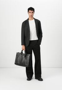 Svart skinn totebag med to topphåndtak, vertikale sømmer og et merkelapp. Bært av en person i en grå blazer og hvit skjorte med svarte bukser.