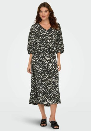 Robe noire avec un motif tacheté blanc, décolleté en V, manches bouffantes, ceinture à la taille, longueur genoux, associée à des sandales noires.