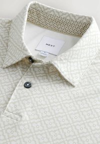 Kortærmet polo-shirt i creme med gentaget geometrisk mønster i lys brun. Har en knap-down krave og en enkelt brystlomme.