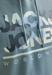 Sudadera verde con un gran texto gráfico "JACK & JONES" en diferentes tonos de azul y blanco. Cuenta con cordones y una textura de tela suave.