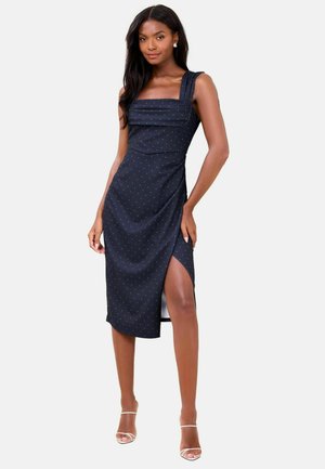 Nő tengerészkék középhosszú ruhában, fehér pöttyökkel, négyzet alakú kivágással, széles pántokkal és magas combon hasított oldallal, fehér magassarkú cipővel párosítva.