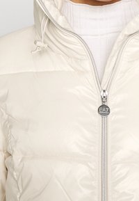 Primo piano di una giacca puffer crema con cappuccio, chiusura frontale con zip e un tirante con il logo EA7 Emporio Armani, indossata sopra un maglione a dolcevita bianco a coste.