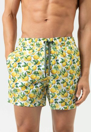 Close-up van een man die witte zwembroek draagt met een gele citroen en groen bladerenpatroon en een grijze trekkoord.