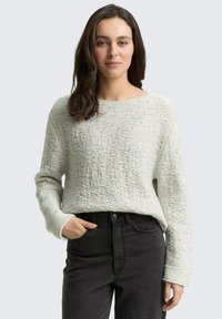 Pull en tricot texturé gris clair avec un décolleté large et des manches longues, associé à un jean noir à taille haute. Matériau doux et tricoté.