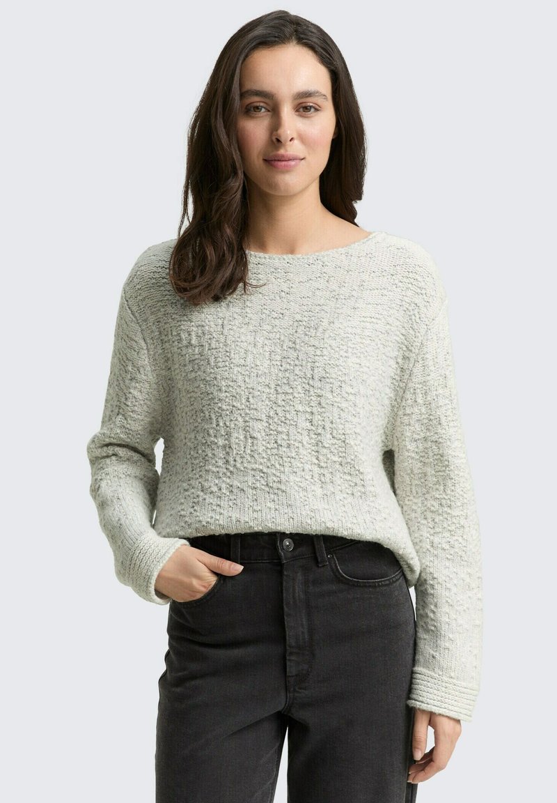 Pull en tricot texturé gris clair avec un décolleté large et des manches longues, associé à un jean noir à taille haute. Matériau doux et tricoté.