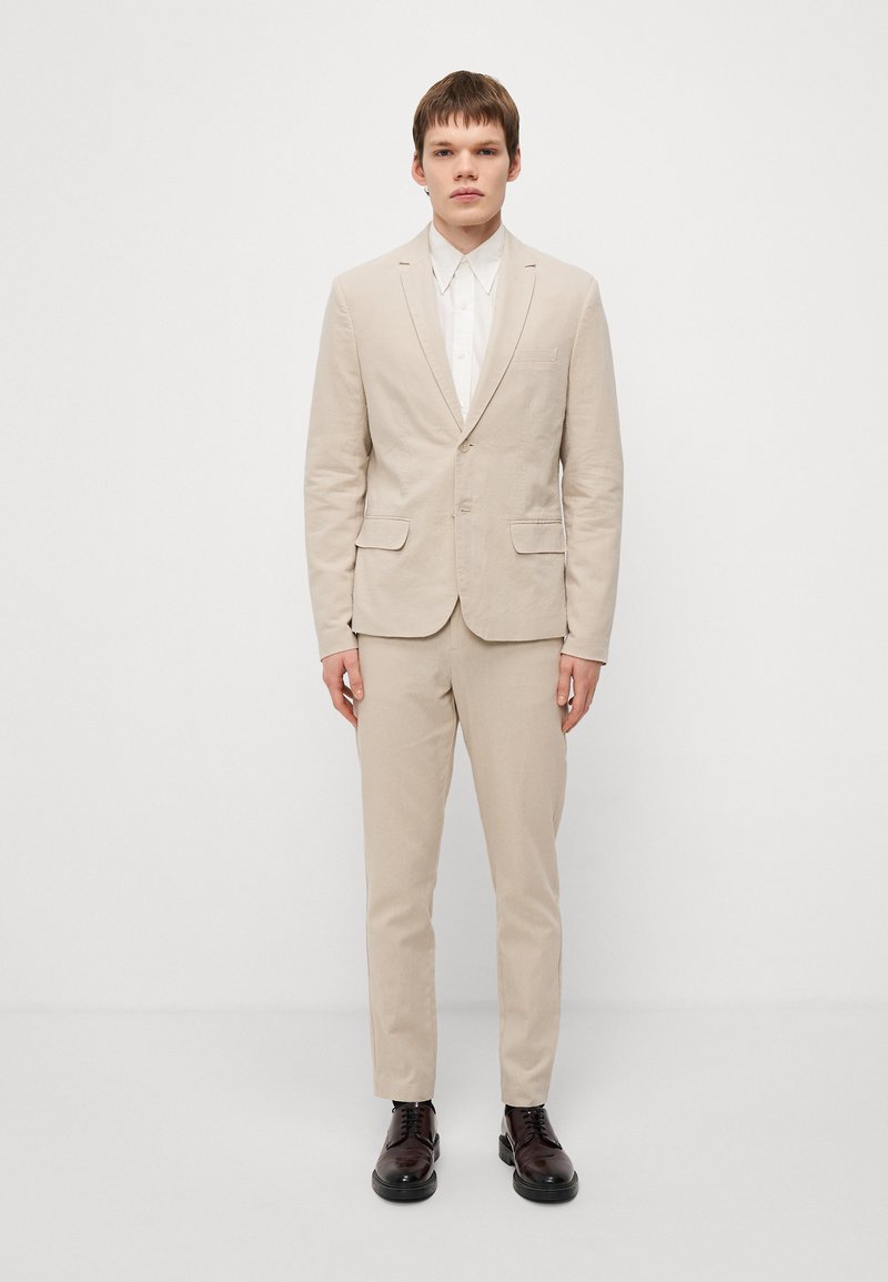 Un costume en lin beige avec deux poches avant, un pantalon assorti et une chemise blanche à col. Des chaussures en cuir marron à lacets complètent la tenue.