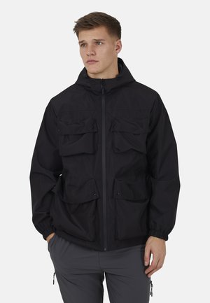 Joven con una chaqueta negra con capucha con cuatro grandes bolsillos delanteros y pantalones grises, de pie con las manos en los bolsillos.