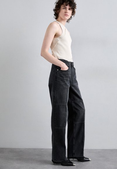 Pantalon large en denim noir avec des détails en patchwork, associé à un débardeur côtelé crème. Le mannequin porte des chaussures noires avec des accents argentés.