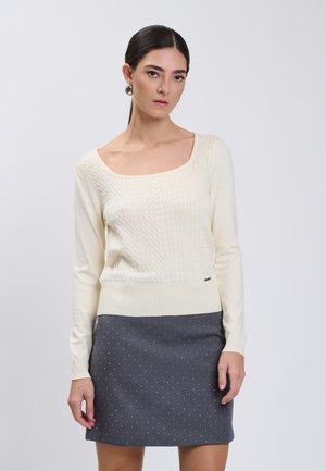 Frau mit dunklem Haar in einem niedrigen Pferdeschwanz, die einen cremefarbenen Zopfpullover und einen grauen Rock mit kleinen weißen Punkten trägt, steht vor einem schlichten Hintergrund.