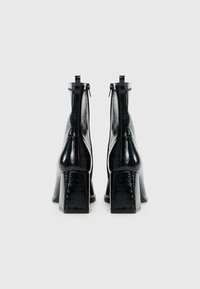 Bottines noires brillantes avec des talons carrés vues de l'arrière, montrant les tirettes de fermeture éclair et la surface en cuir texturé.