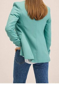 Un blazer verde menta su misura presenta spalle strutturate, uno spacco posteriore e un design slanciato abbinato a jeans blu casual.