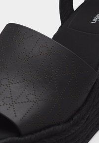 Sandale noire à bout ouvert avec un motif étoile perforé sur la bride supérieure et une semelle tissée texturée.