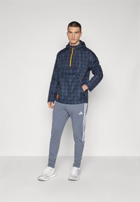 Chaqueta de pullover de patrón navy con un bolsillo frontal y cremallera amarilla, combinada con pantalones de chándal grises que cuentan con franjas blancas y un logo.