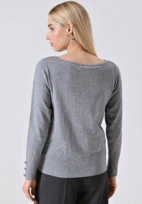 Maglione grigio leggero con maniche lunghe, scollatura ampia e polsini con bottoni. Tessuto morbido e design aderente, ideale per essere indossato a strati.