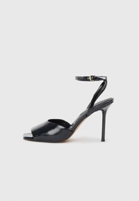 Sandal de charol negro con tacón alto, punta abierta, tacón stilettos delgado y correa al tobillo con hebilla dorada sobre fondo blanco.