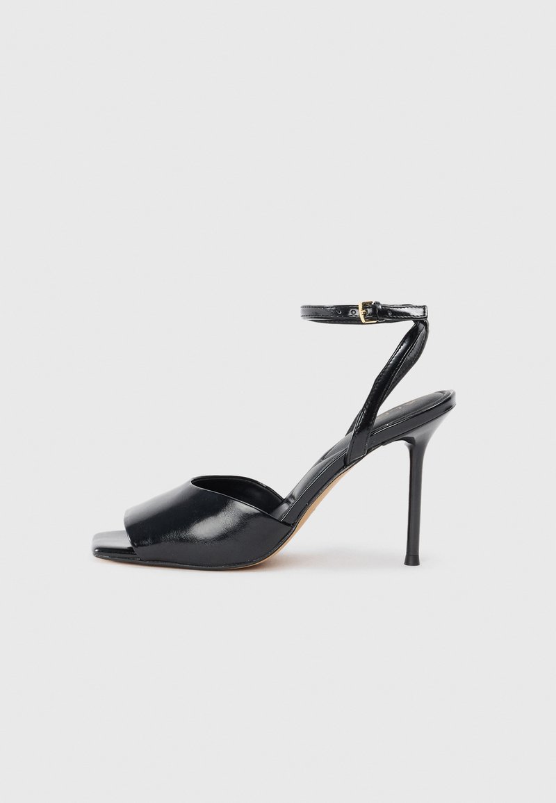 Sandal de charol negro con tacón alto, punta abierta, tacón stilettos delgado y correa al tobillo con hebilla dorada sobre fondo blanco.