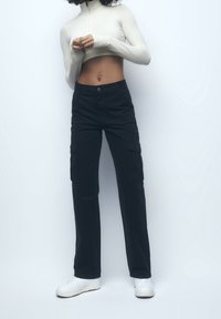 Personne se tenant contre un mur, portant un haut blanc à manches longues court, un pantalon cargo noir et des baskets blanches, les mains jointes devant.