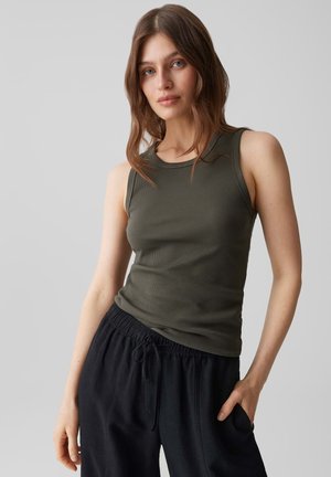 Jonge vrouw met bruin haar die een mouwloze, olijfgroene, geribbelde top en losse zwarte broek draagt, staande met één hand in de zak.