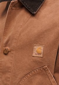 Καφέ μ πουφάν από καμβά με γιακά από κοτλέ, κλείσιμο με κουμπιά και τετράγωνη ετικέτα με κίτρινο λογότυπο και όνομα μάρκας.