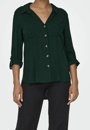 Mujer con camisa abotonada de color verde oscuro con las mangas remangadas y pantalones negros, de pie con los brazos relajados a los lados.