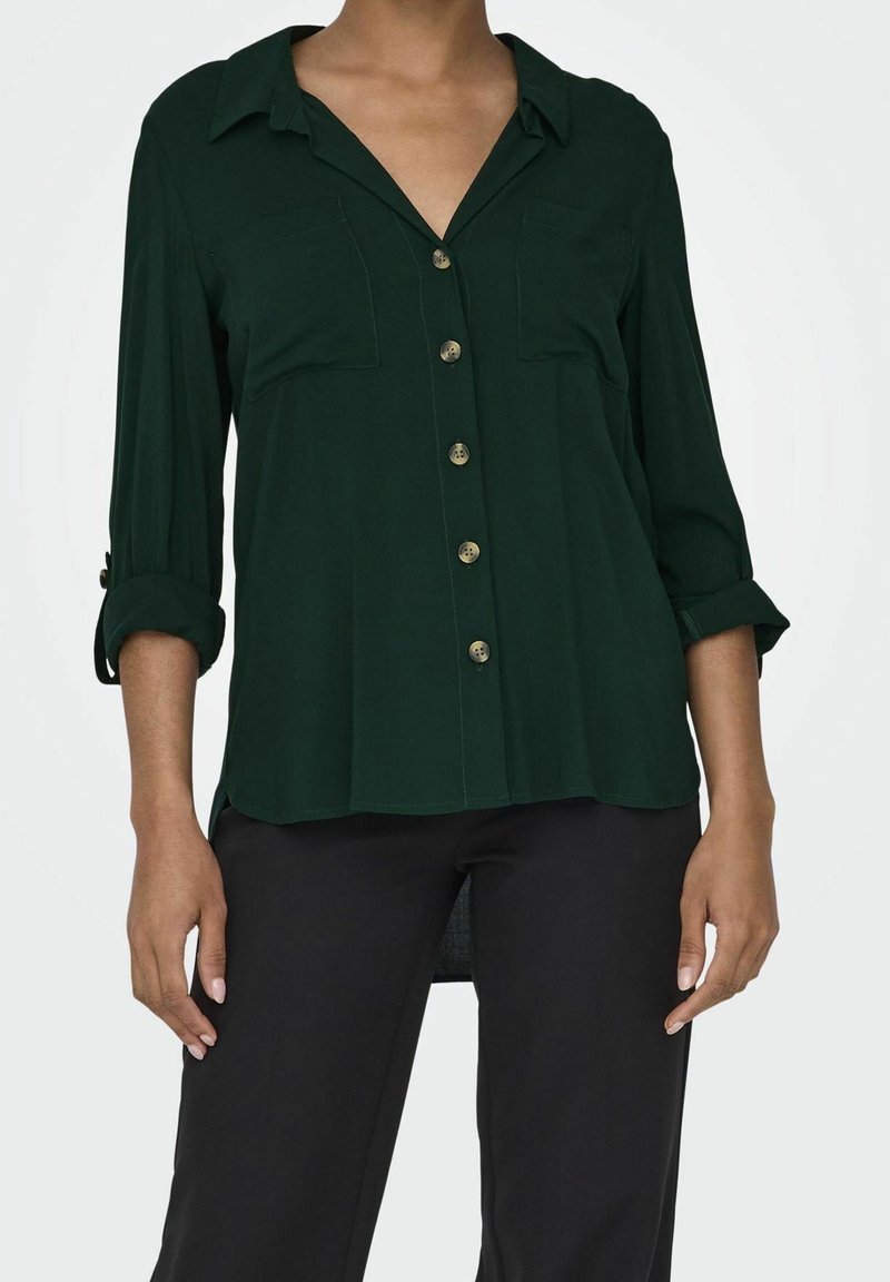 Femme portant une chemise boutonnée vert foncé avec les manches retroussées et un pantalon noir, debout avec les bras détendus le long du corps.