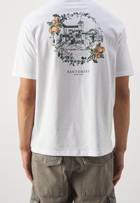 Bílé bavlněné tričko s grafikou Santorini a kulatým květinovým a oranžovým designem na zádech. Volný střih, krátké rukávy.