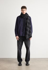 Fular negru cu un model repetat "HUGO", purtat cu o cămașă button-up de culoare purpurie închisă și blugi negri, stând pe un podea din beton deschis.