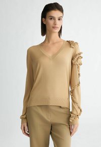 Maglione beige a maniche lunghe con scollo a V, caratterizzato da dettagli arricciati lungo una spalla e una manica, e una vestibilità comoda.