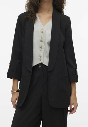 Frau trägt schwarzen Blazer mit hochgekrempelten Ärmeln, schwarze Hose und hellgraues ärmelloses, geknöpftes Oberteil, steht vor weißem Hintergrund.