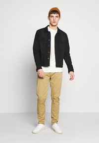 Črno denim jakno, belo majico, bež chinos hlače in bele superge. Model nosi oranžno pleteno kapo in stoji proti nevtralni ozadju.
