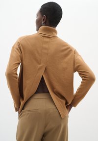 Brun rullekrave-trøje med en åben detalje i midten af ryggen, lavet af blødt stof. Kombineret med beige bukser med en afslappet pasform.