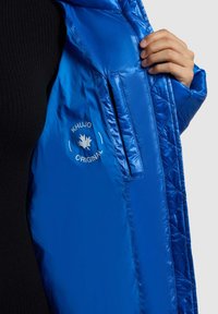 Blaue Steppjacke mit einem gesteppte Design, glatter, glanzender Textur und einem Logo-Patch, mit einer verborgenen Seitentasche innen.