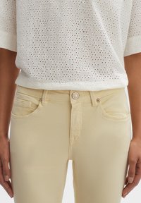 Jean en denim jaune clair avec une coupe ajust ée, cinq poches et une fermeture à bouton rond. Accompagné d'un chemisier blanc en broderie anglaise.