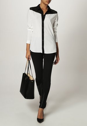 Mujer vestida con una blusa blanca con canesú de encaje negro, pantalones negros con paneles inferiores texturizados, tacones negros y llevando un bolso negro.