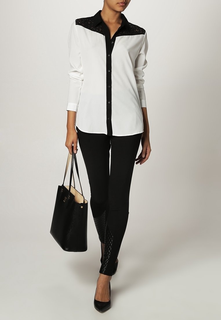 Femme portant un chemisier blanc avec un empiècement en dentelle noire, pantalon noir avec des panneaux inférieurs texturés, talons noirs, tenant un sac à main noir.