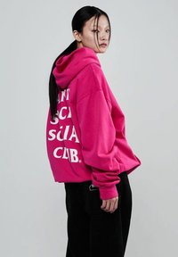 Modelka z długimi czarnymi włosami nosi jasnoróżową, oversize'ową bluzę z odważnym białym napisem, zestawioną z czarnymi spodniami, na jasnym tle.