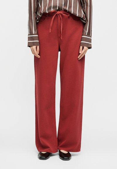 Ευρύχωρο κόκκινο παντελόνι με κορδόνι στη μέση, συνδυασμένο με καφέ και λευκή ριγέ μακρυμάνικη μπλούζα και μαύρα παπούτσια με τετράγωνη μύτη.