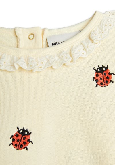 Pull crème avec un col rufflé et des coccinelles rouges brodées sur le devant ; le tissu a une finition tricotée texturée.