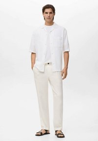 Camicia leggera bianca a maniche corte, colletto aperto, taschino frontale; pantaloni color crema; sandali neri. Tessuto morbido, vestibilità comoda.