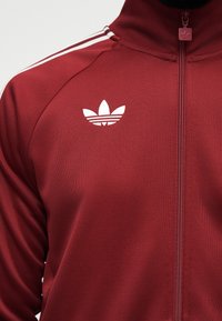 Vinrød sportjakke med lynlås, med hvid Adidas trefoil-logo på brystet og hvide striber langs skuldersømmen.